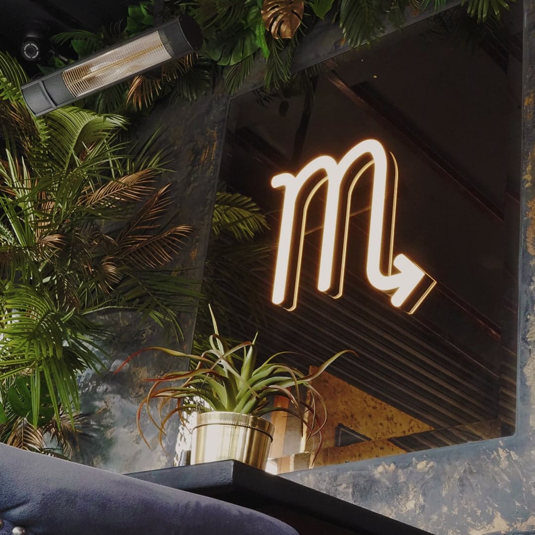 A neon sign glowing above a banquette, palms and a brass planter
